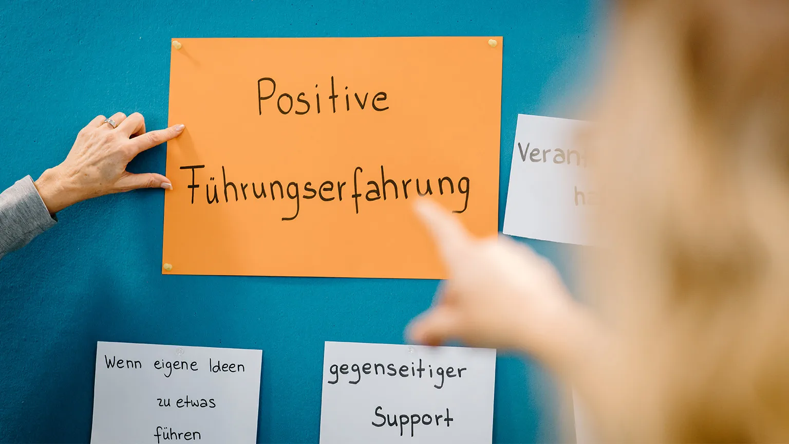 Hände zeigen auf einen Zettel, der zusammen mit anderen Zetteln an einer Pinnwand hängt. Auf der Zettel steht positive Fürungserfahrung.
