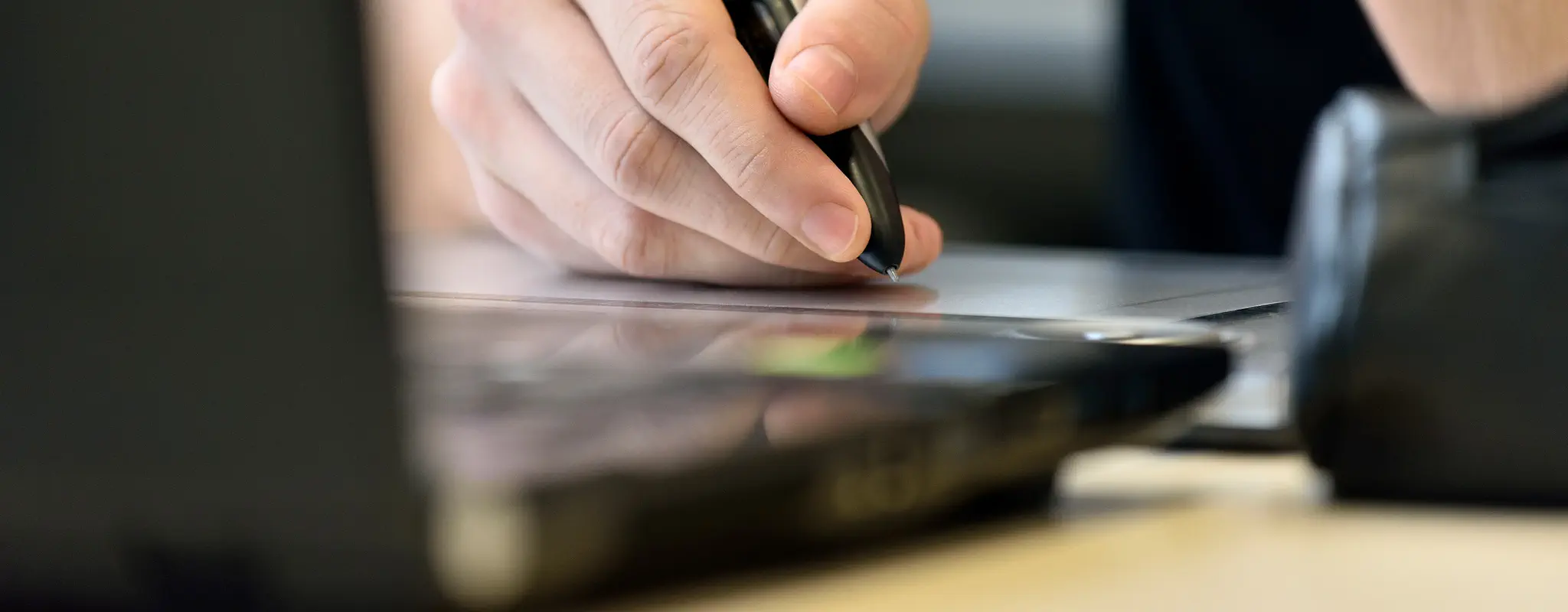 Hand mit einem schwarzen Kugelschreiber an einem Grafiktablet, davor steht ein Notebook.