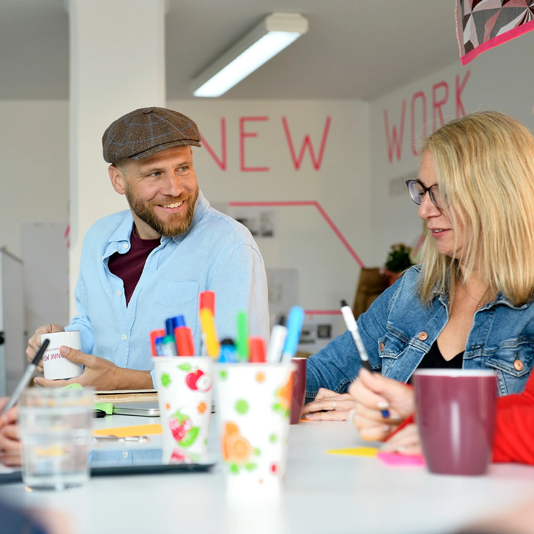 Mann und Frau sitzen lächelnd an einem Tisch, auf dem bunte Becher mit Stiften und weitere Arbeitsmaterialien liegen