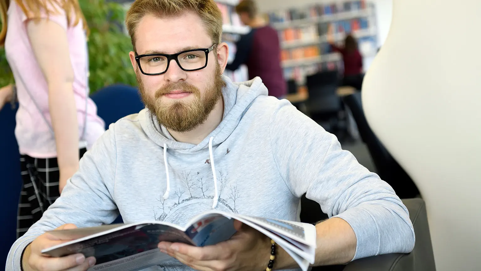 Ein Student sitz in der Bibliothek der HSW und hält ein Buch in der Hand während er in die Kamera lächelt.