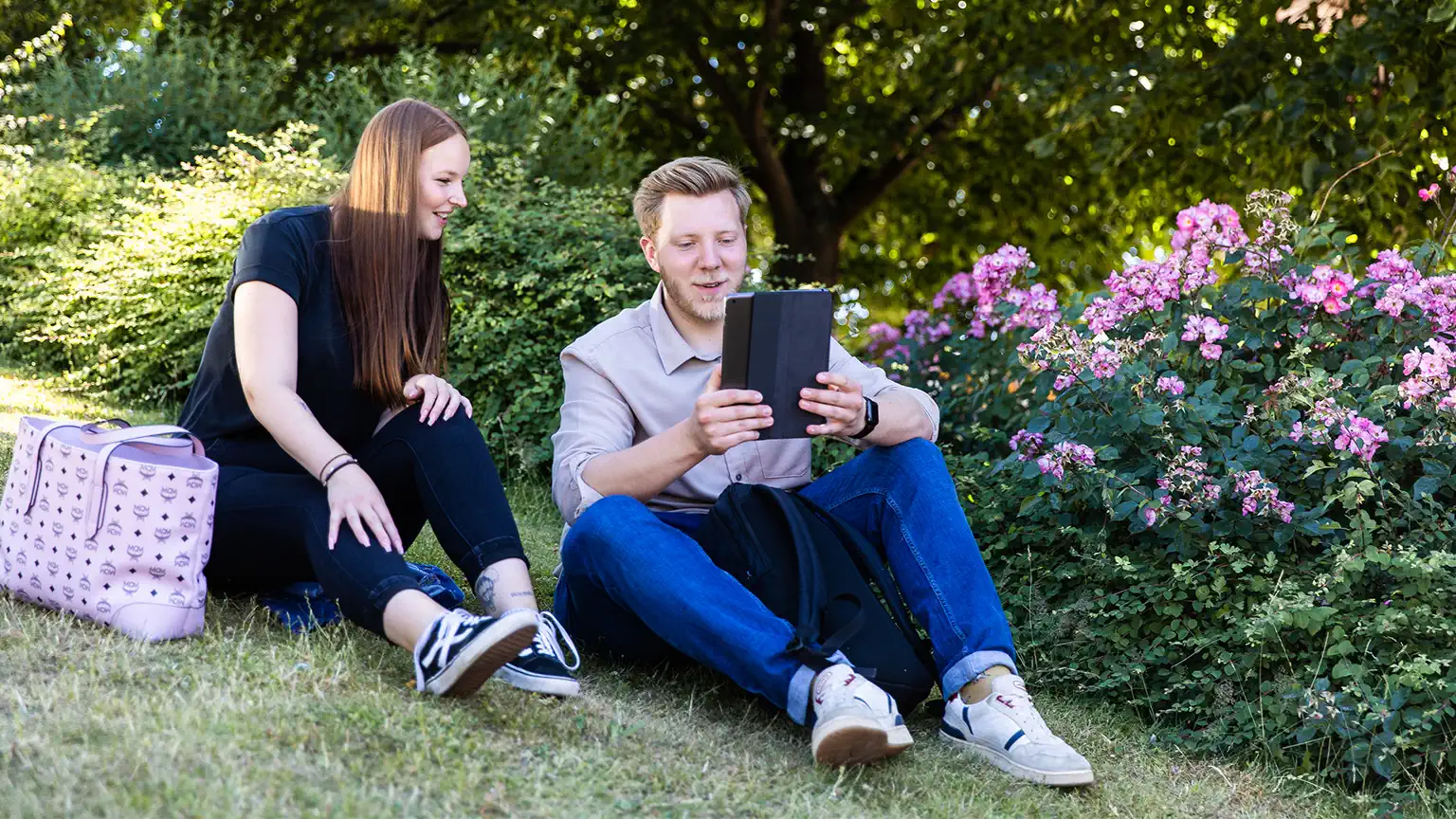 Ein Student und eine Studentin sitzen auf einer Wiese. Er zeigt ihr lachend etwas auf einem Tablet.