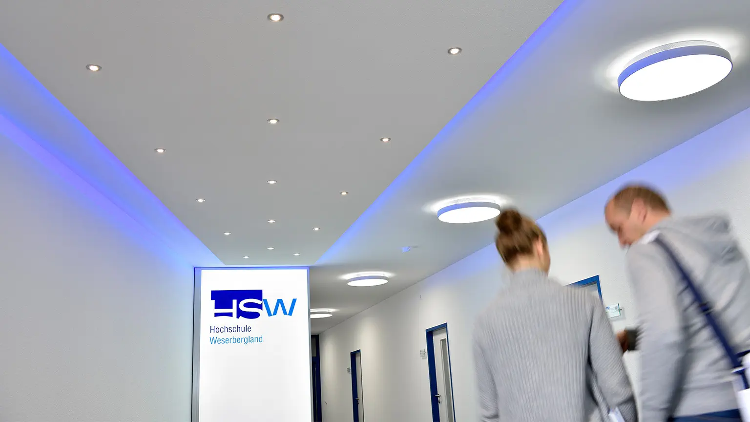 Eine Frau und ein Mann laufen auf dem Flur der HSW. Rechts von ihnen sieht man Türen mit blauem Rahmen und links ein beleuchtetes Schild mit dem Logo der HSW sowie Spotlights an der decke, die leuchten.