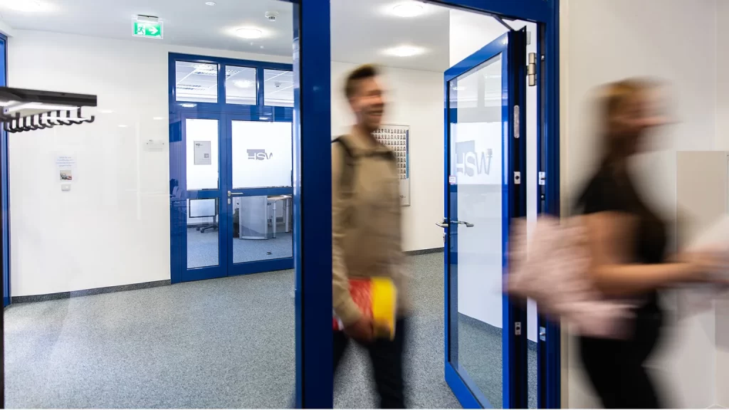Zwei Studierende laufen in einem Flur durch eine blaue Glastür mit der Aufschrift HSW. Im Hintergrund ist eine weitere Glastür zu erkennen.