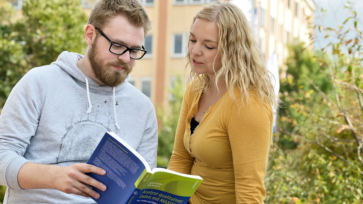 Ein Student und eine Studierende sitzen draußen vor dem Gebäude der HSW und schauen gemeinsam in ein Buch, das der Student in seinen Händen hält.