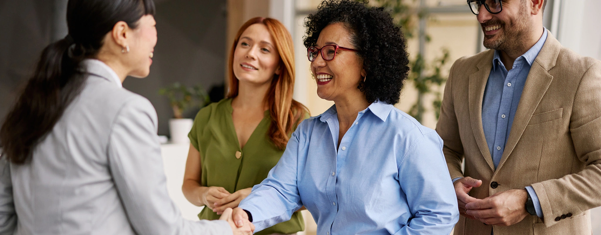 Vier lächelnde Personen mit internationaler Herkunft stehen zusammen, eine Frau reicht einer anderen die Hand