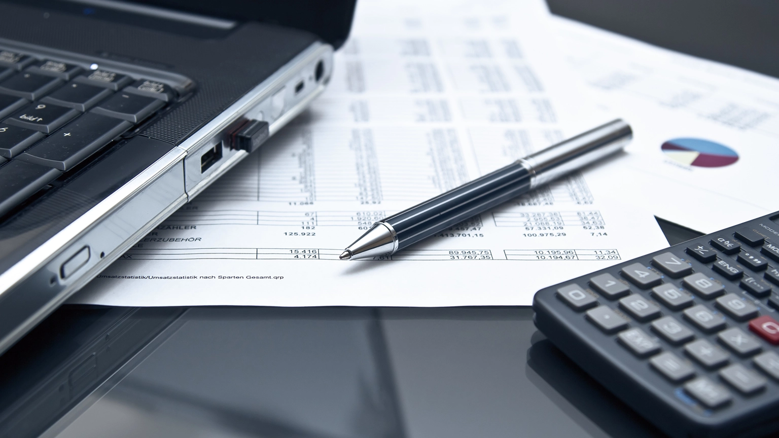 Stift und Taschenrechner liegen mit Papieren auf einem tisch und vor einem Laptop