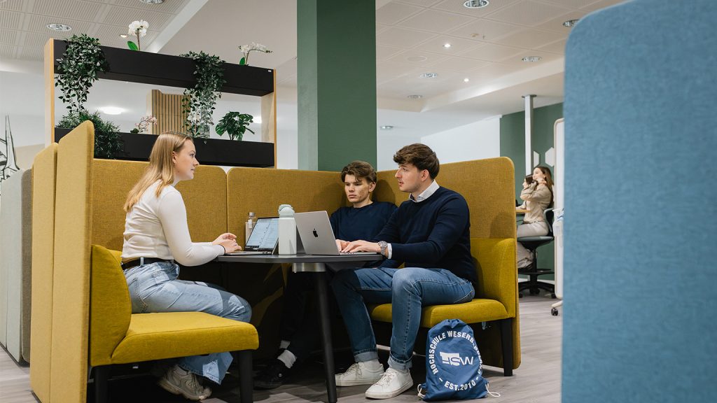 Drei Studierende sitzen in einer gelben Sitzecke mit Laptop auf dem Tisch