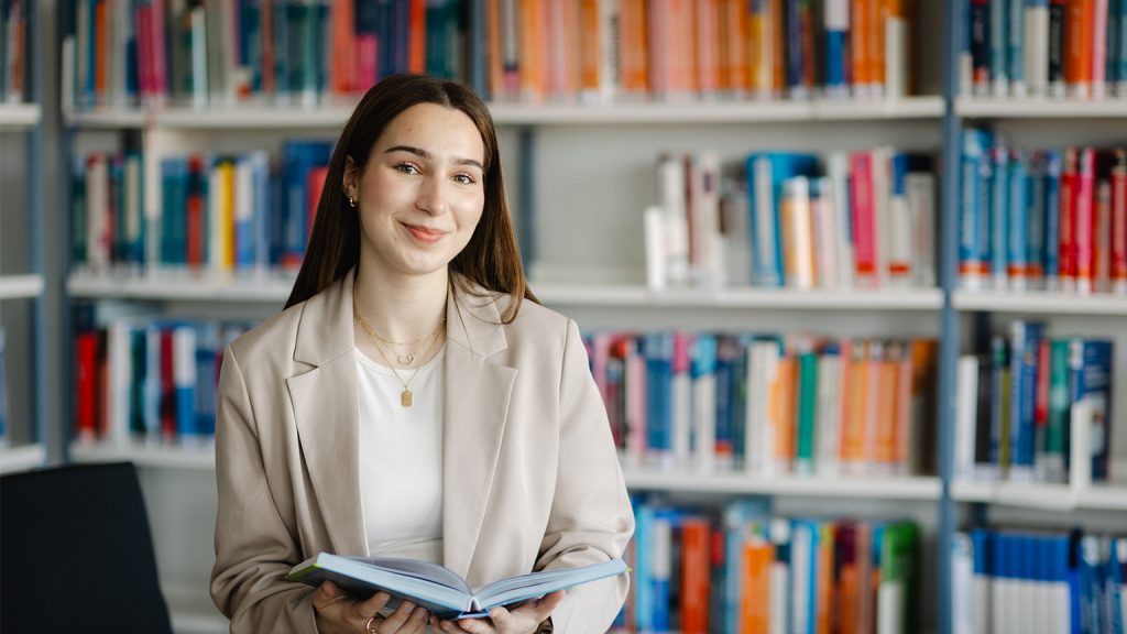 Eine Damen hält ein aufgeschlagenes Buch in der Hand. Hinter ihr ist eine Buchwand zu sehen.