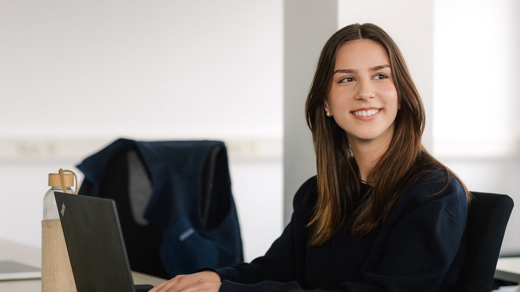 Eine Studierende mit Laptop vor sich lächelt in die Kamera