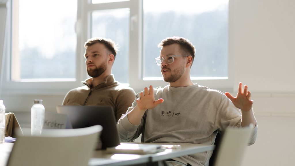 Zwei jungen Männer sitzen am Tisch nebeneinander. Einer gestikuliert mit den Händen.